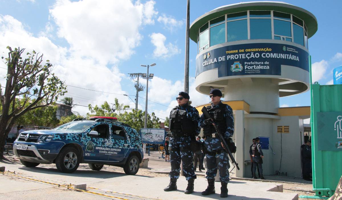 torre de segurança ao fundo e dois guardad municipais posando em pé na frente e viaturas ao lado